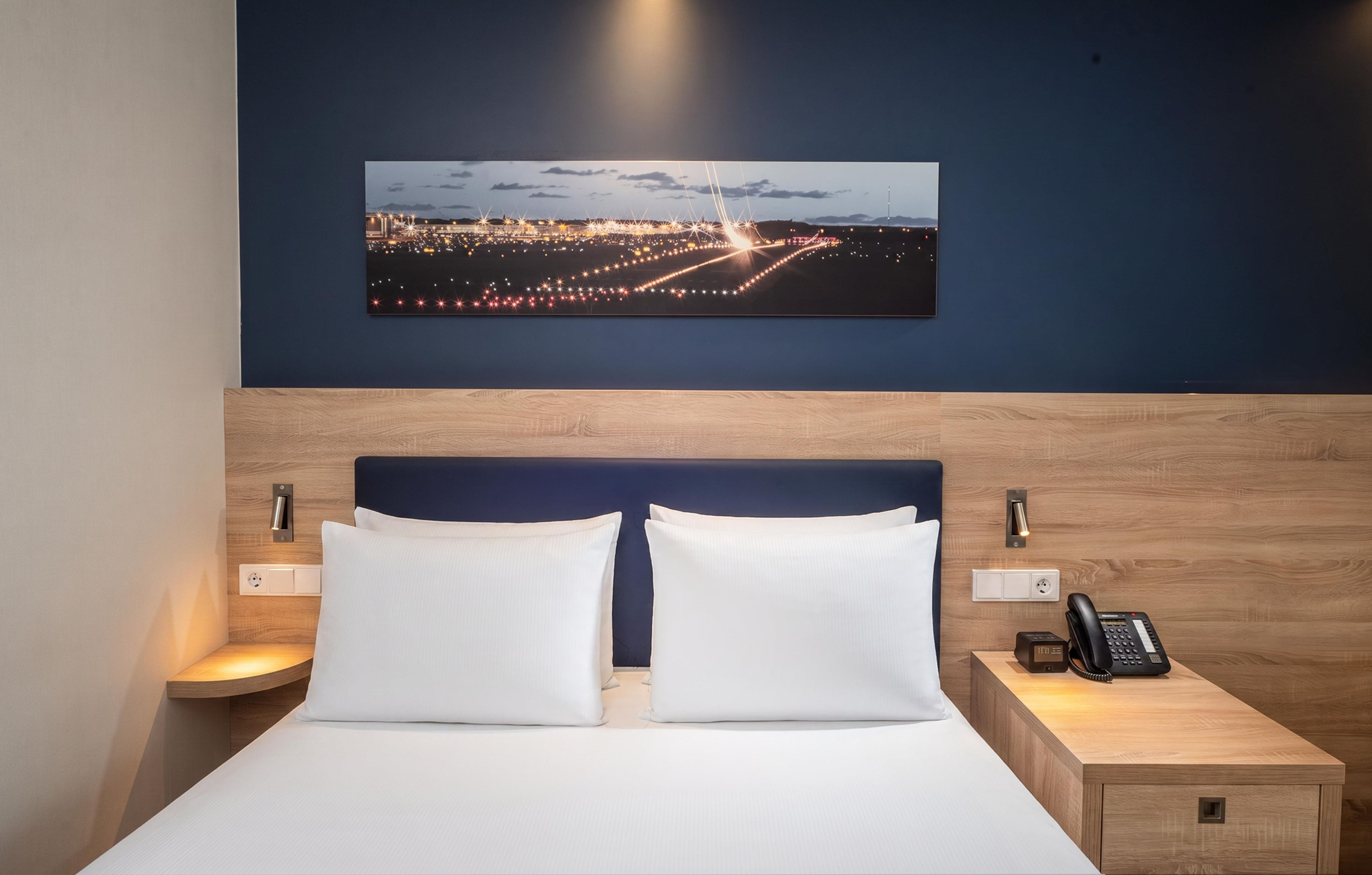 A hotel room with a bed, two bedside tables, a nightstand, a wall-mounted phone, and a cityscape painting above the bed.