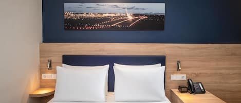 A hotel room with a bed, two bedside tables, a nightstand, a wall-mounted phone, and a cityscape painting above the bed.