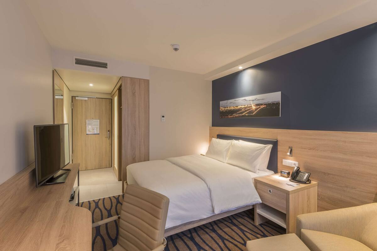 Ein Hotelzimmer mit Bett, Nachttisch, Sessel, Fernseher und einem Bild an der Wand.