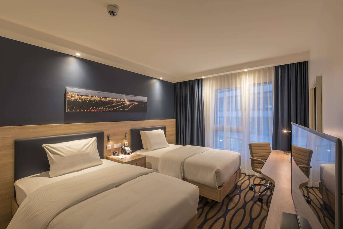 Ein Hotelzimmer mit zwei Betten, einem Schreibtisch, einem Stuhl, einem großen Fenster mit Vorhängen, einem Stadtbild an der Wand und einer Deckenleuchte.