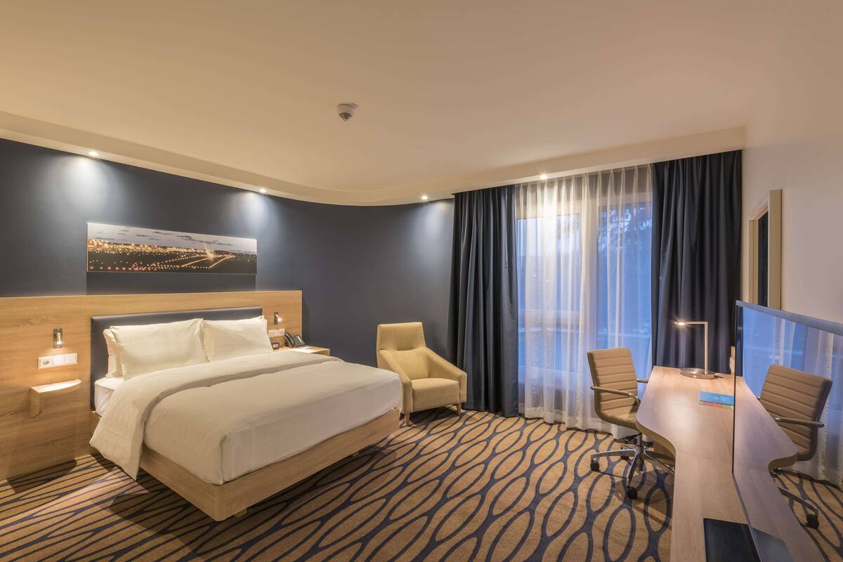 Ein Hotelzimmer mit einem großen Bett, einem Schreibtisch mit zwei Stühlen, einem Sessel, einem Nachttisch und einem Gemälde an der Wand.