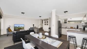 A modern apartment with a dining area, living room, kitchen, and a TV displaying a cityscape.