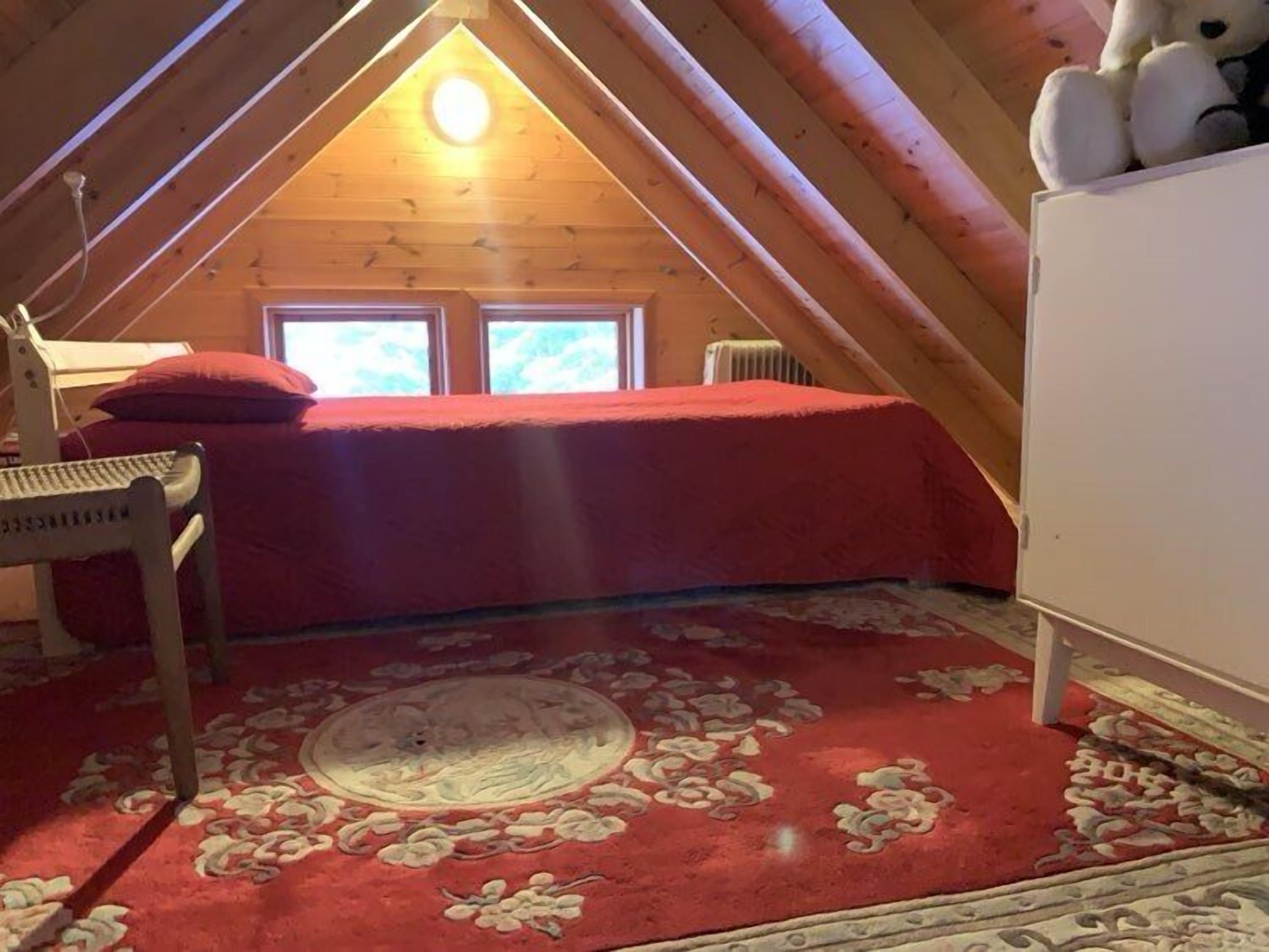 A cozy attic bedroom with a red bedspread, a wooden chair, and a patterned carpet.