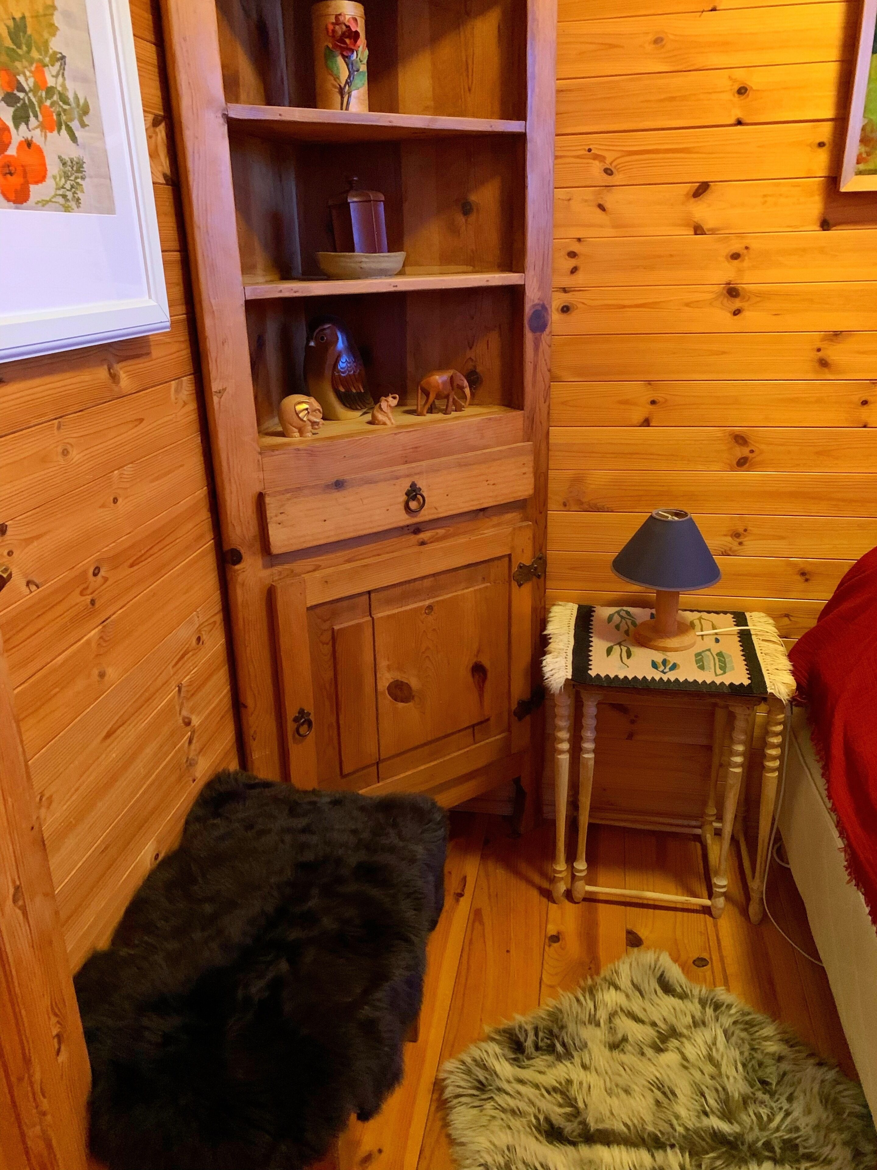 A wooden room with a shelf containing decorative items, a lamp on a small table, and a fur rug on the floor.
