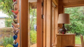 Un interno in legno con una porta che dà su un giardino, una lampada su un tavolo di legno e una mensola con dei libri.
