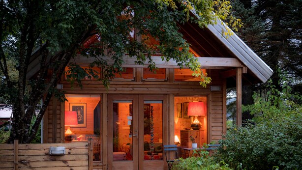 Una baita di legno con ingresso a porta in vetro, un piccolo portico e una recinzione in legno.