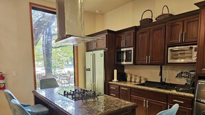 A kitchen with dark wood cabinets, a granite countertop, a gas stove, a refrigerator, a microwave, and a dishwasher. There are two bar stools and a window with a view of trees.