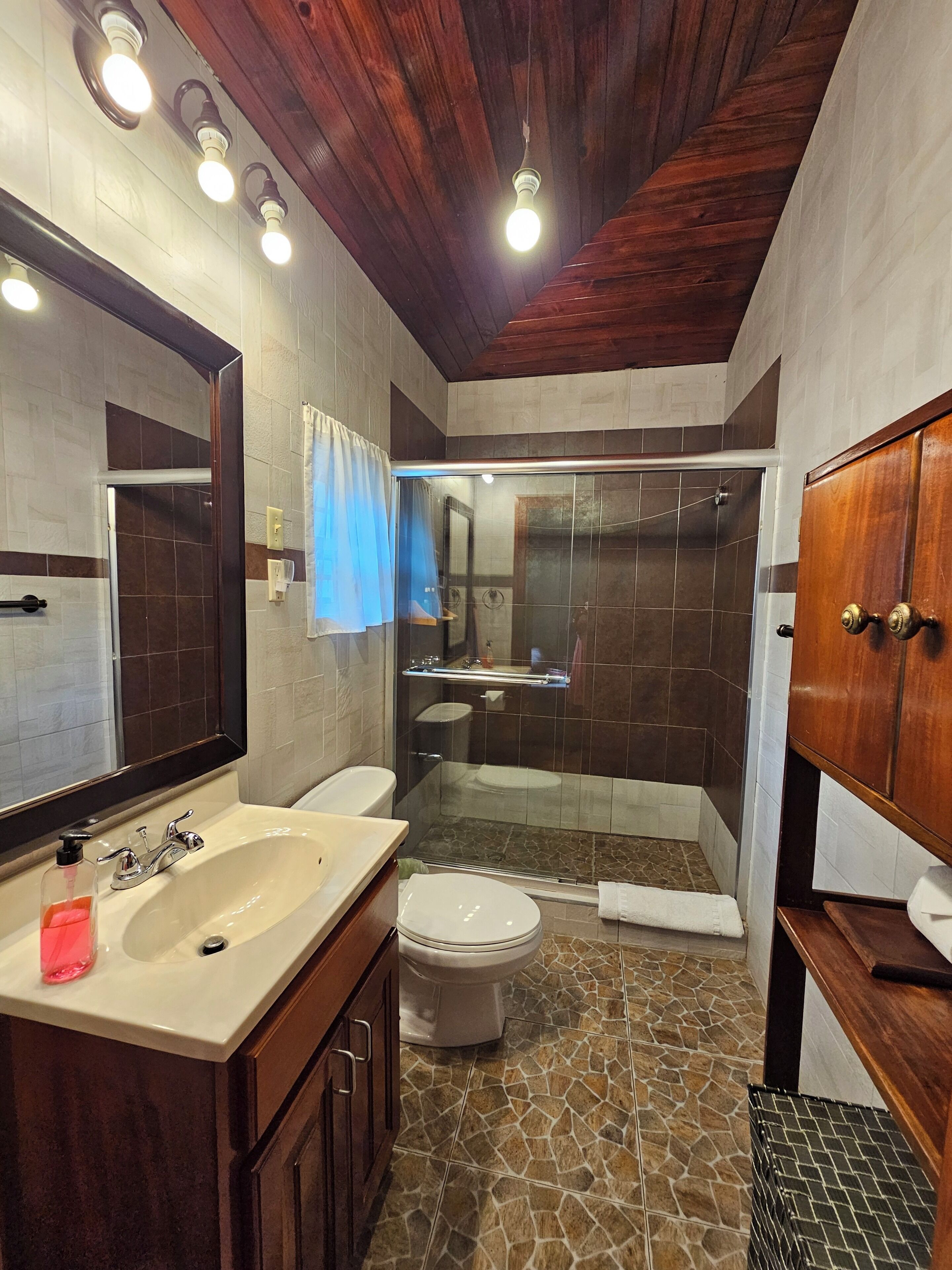 A bathroom with a double sink, a toilet, a shower with glass doors, and a wooden ceiling.