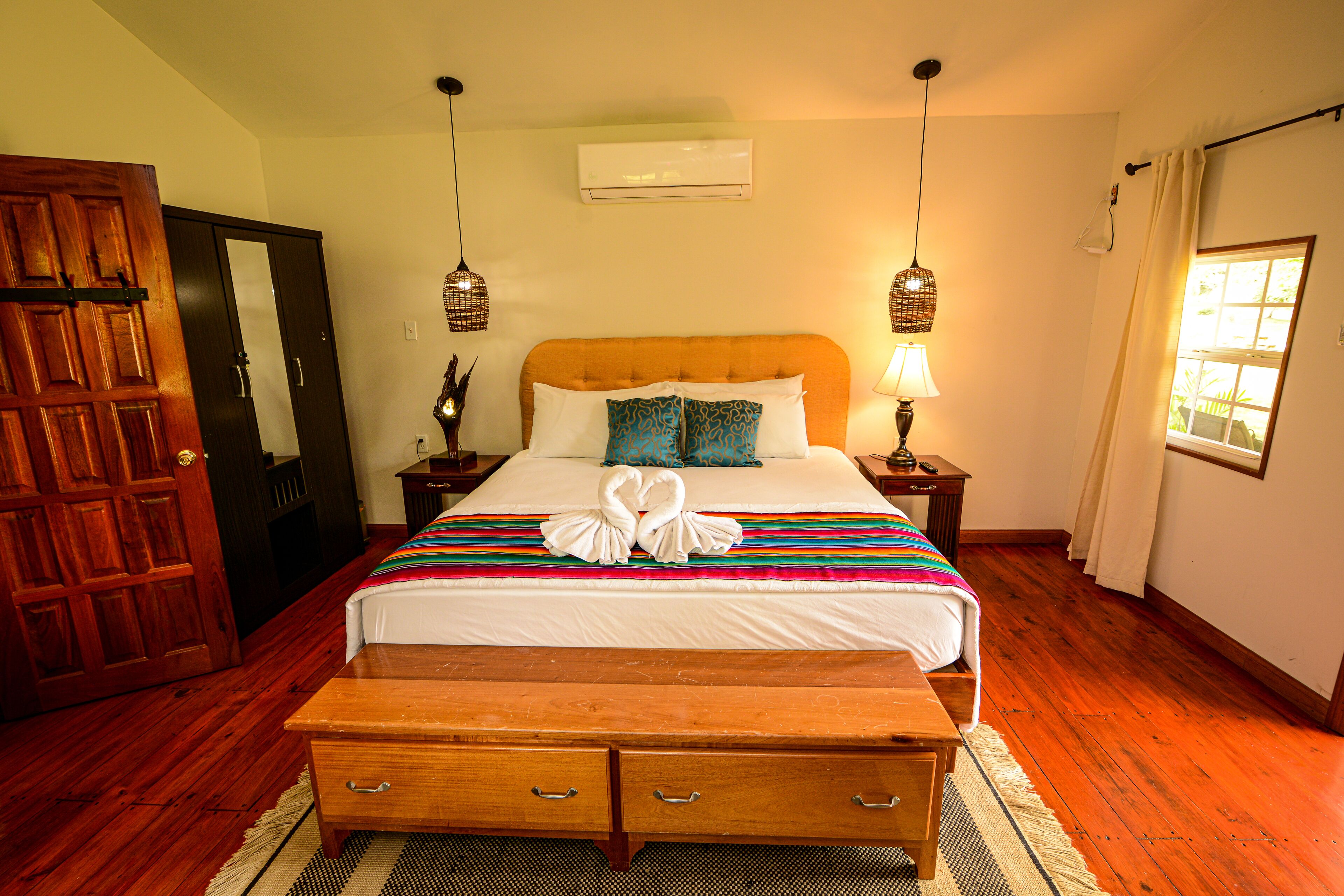 Quarto de hotel com cama de madeira, um banco, mesa de cabeceira, ar condicionado e porta de madeira.