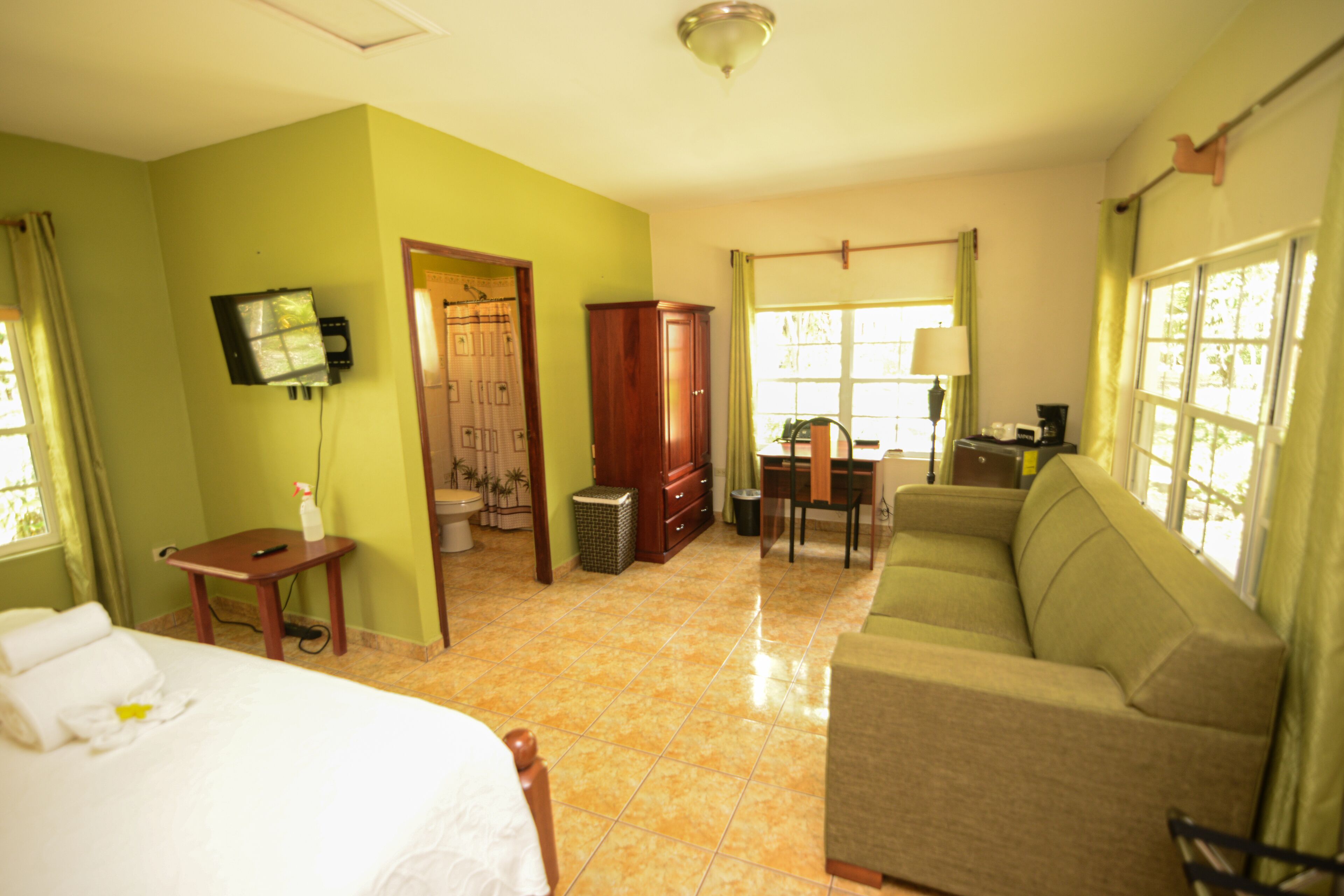 A hotel room with a bed, a sofa, a desk with a chair, a television mounted on the wall, and a window with curtains.