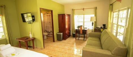 A hotel room with a bed, a sofa, a desk with a chair, a television mounted on the wall, and a window with curtains.