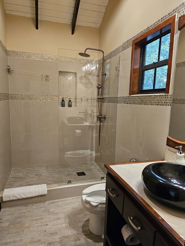 A modern bathroom with a glass-enclosed shower, a freestanding bathtub, a vanity with a round black sink, and a window with a view of trees.