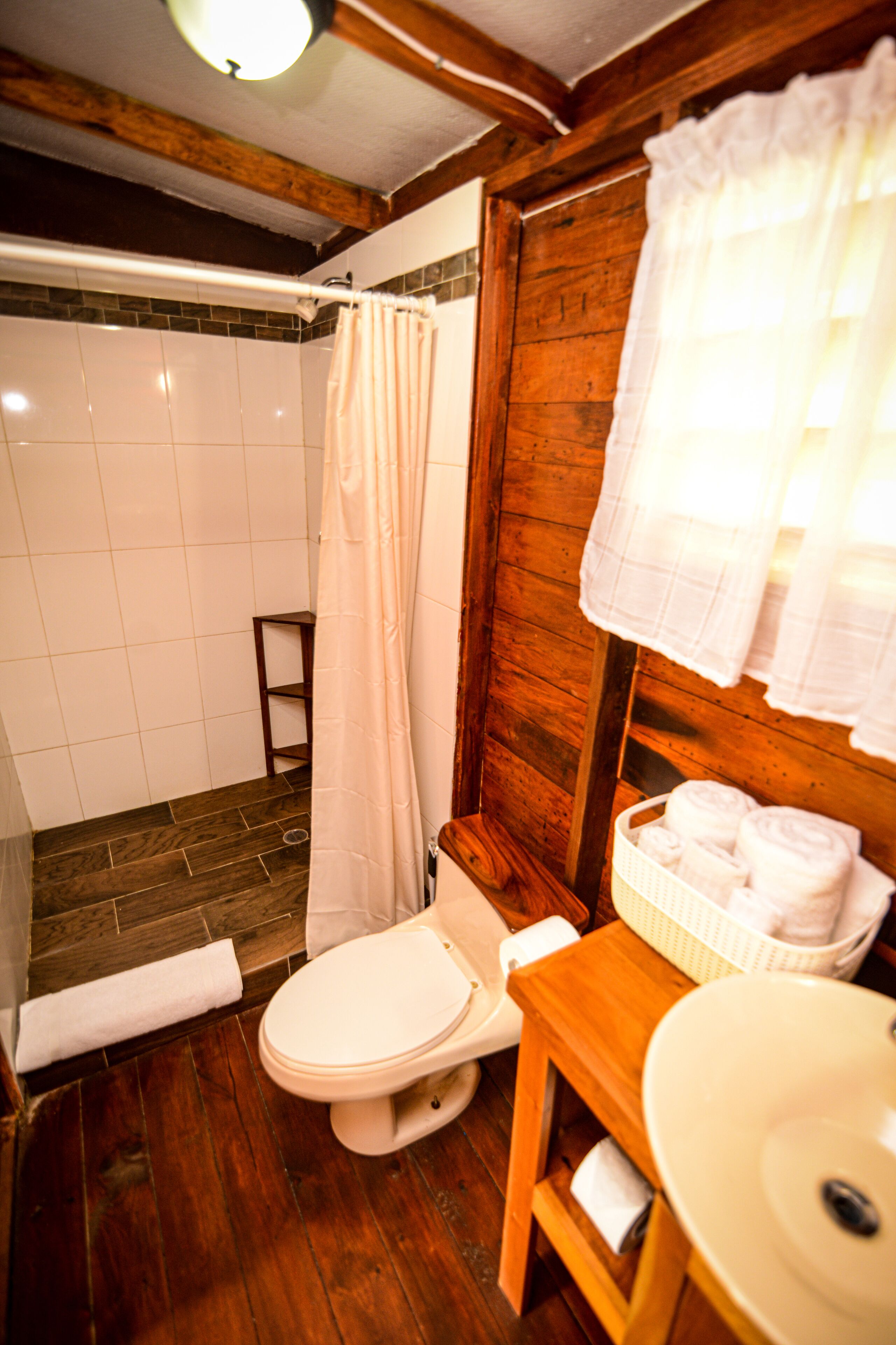 A bathroom with a wooden floor, a white toilet, a wooden sink, a white bathtub with a shower curtain, and a wooden shelf with towels.