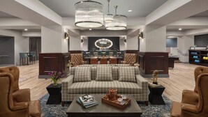A spacious lobby with a checkered sofa, leather armchairs, a coffee table with magazines and a tray, and a dining area in the background. - Hotel Northland, Autograph Collection (Green Bay)