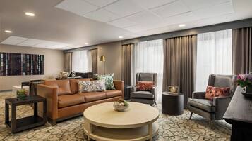 A modern hotel room with a brown leather sofa, two armchairs, a round coffee table, and a large window with curtains.