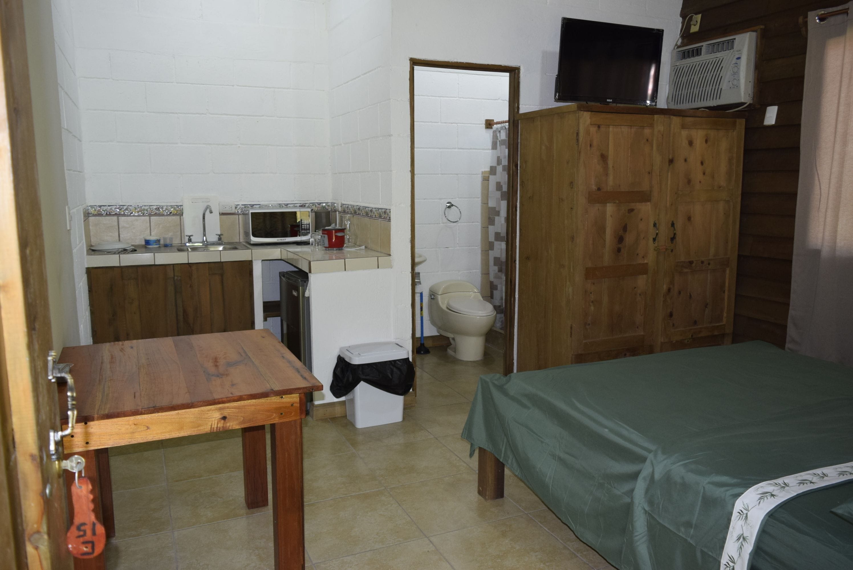 Una habitación compacta con una mesa de madera, una cama, una pequeña cocina con microondas y un inodoro.
