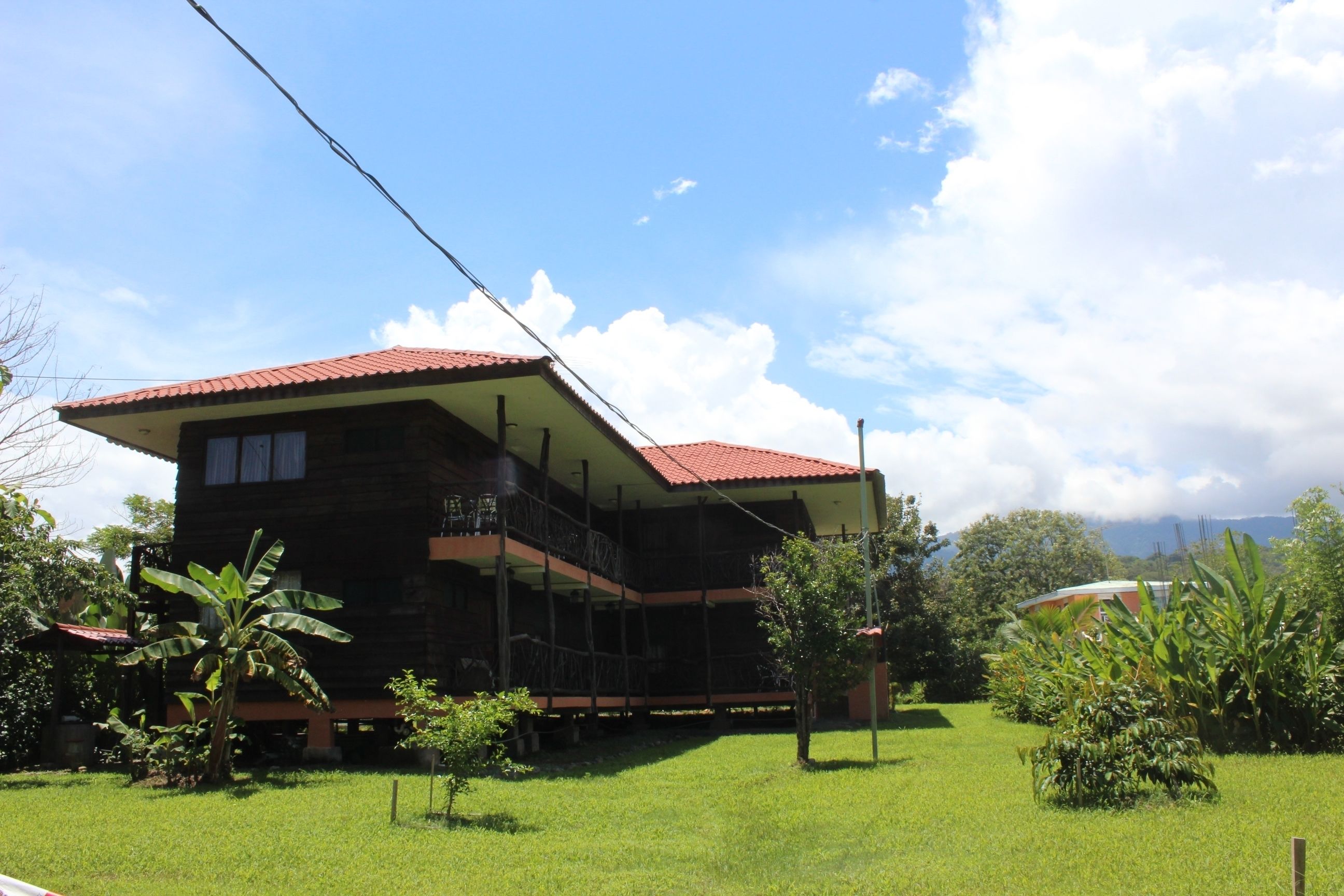 A two-story wooden house with a red roof, a balcony on the upper floor, and a grassy yard.