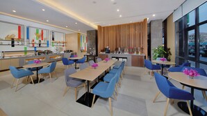 A modern dining area with multiple tables and chairs, a kitchen area with appliances, and large windows offering a view. - Holiday Inn Express Zhangjiagang East by IHG (Suzhou)