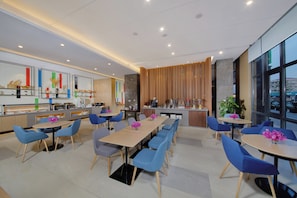 A modern dining area with multiple tables and chairs, a kitchen area with appliances, and large windows offering a view. - Holiday Inn Express Zhangjiagang East by IHG (Suzhou)