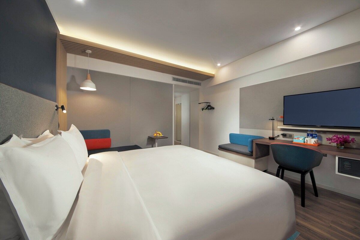 A hotel room with a bed, a desk, a chair, a television, and a small table with a lamp.