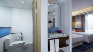 A modern hotel bathroom with a toilet, sink, and mirror. There is a bed with a blue headboard and a window with curtains. - Holiday Inn Express Zhangjiagang East by IHG (Suzhou)