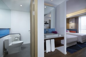 A modern hotel bathroom with a toilet, sink, and mirror. There is a bed with a blue headboard and a window with curtains.