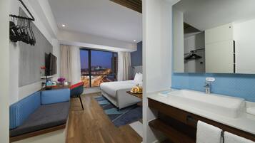 Quarto de hotel moderno com uma cama grande, área de estar, banheiro com pia e vista para a cidade.