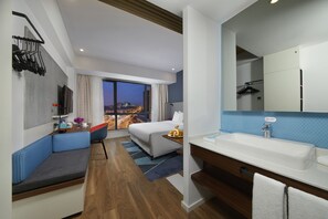 A modern hotel room with a large bed, a seating area, a bathroom with a sink, and a city view.