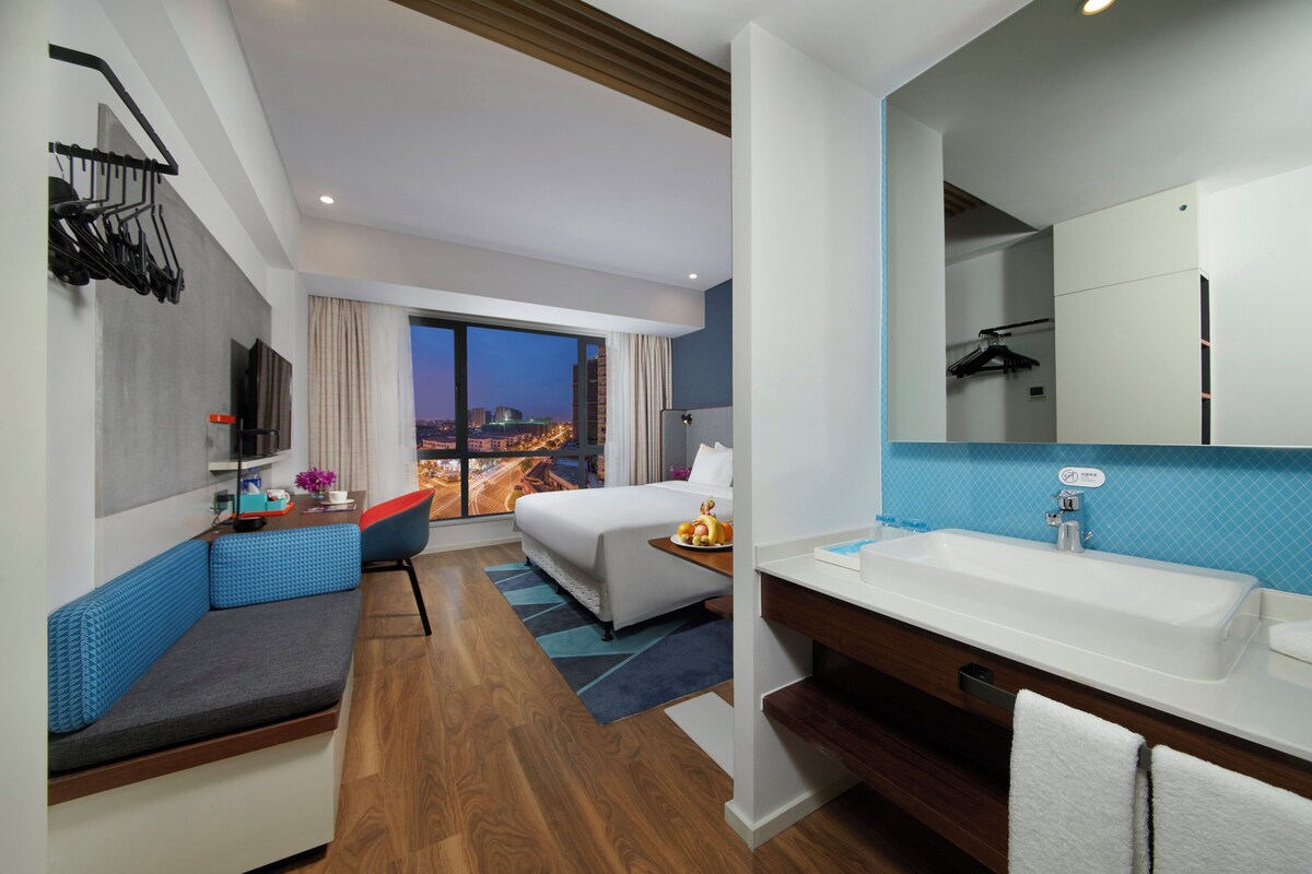 A modern hotel room with a bed, seating area, and a large bathroom with a sink and mirror.
