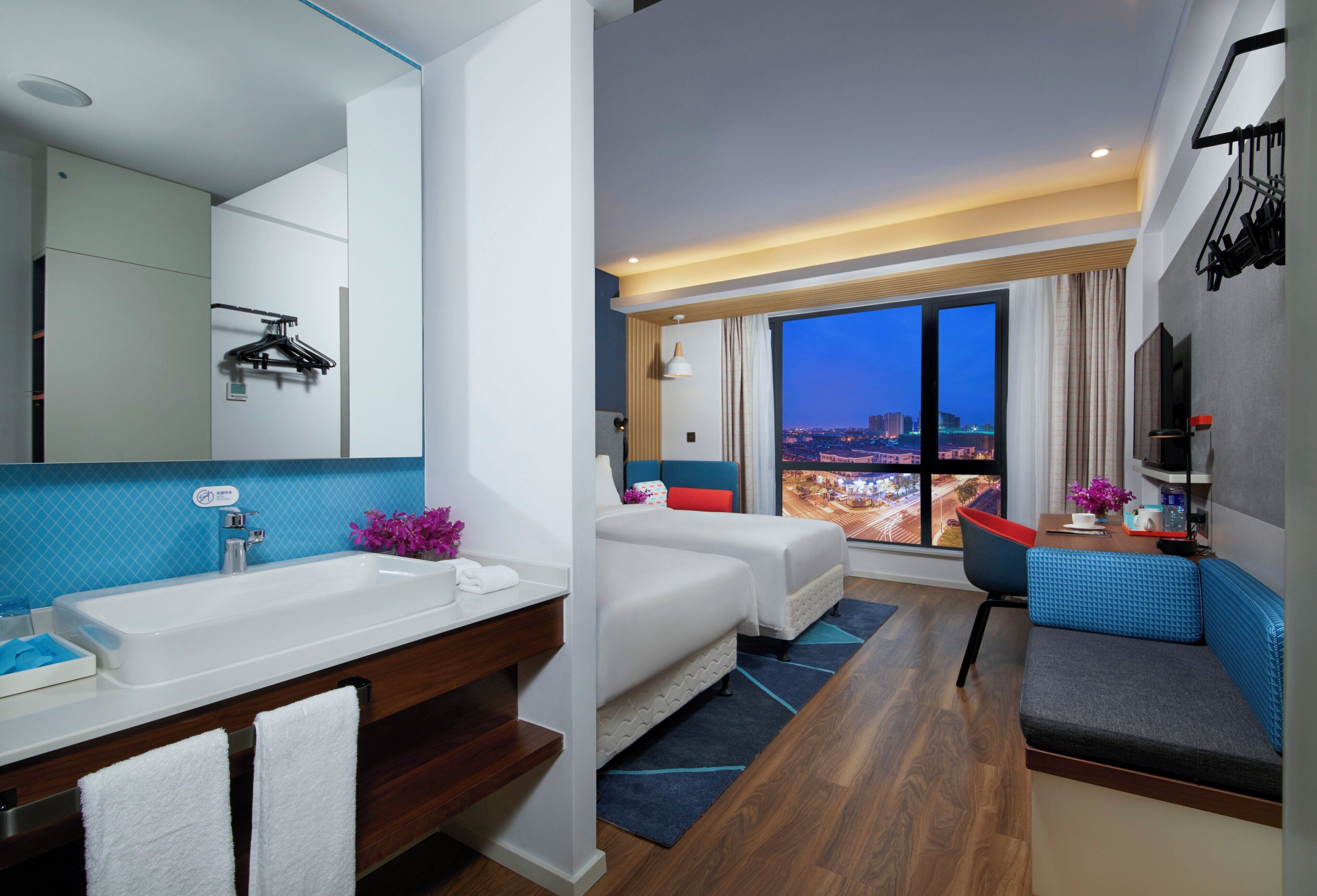 A modern hotel room with a large window, a bathroom with a sink and mirror, and a seating area with a view of the city.