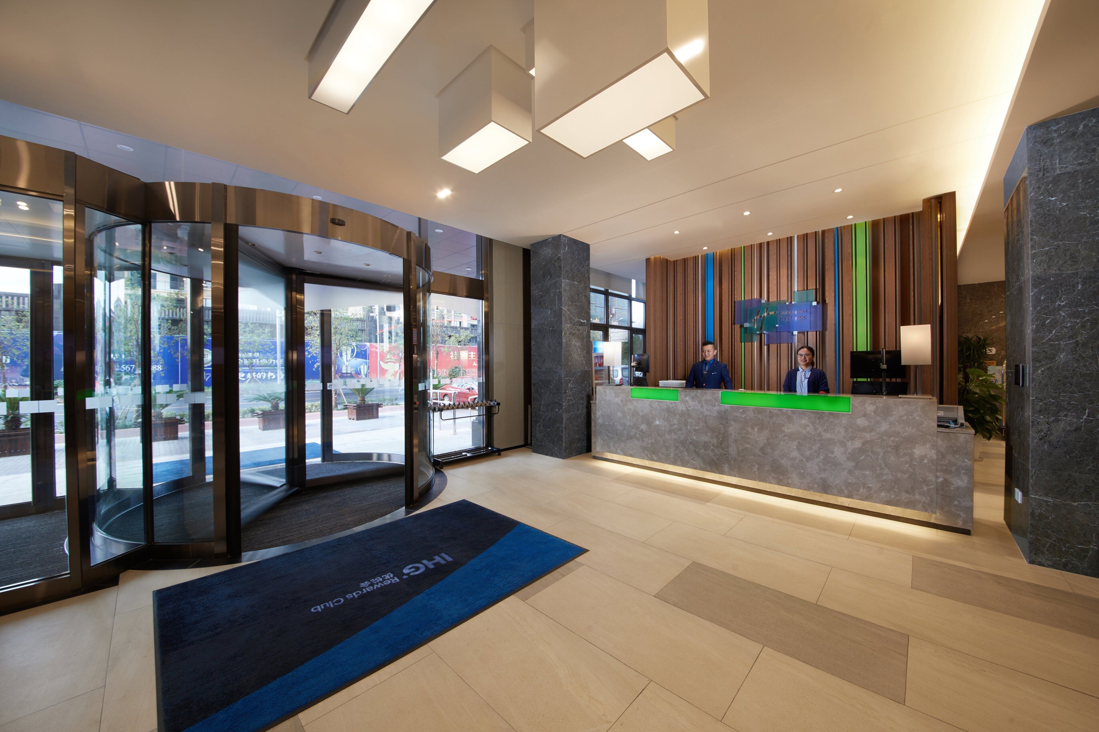 Área de recepção de hotel com design moderno, incluindo uma porta giratória de vidro, um balcão de atendimento e vista para a rua.