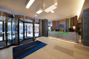 Área de recepção de hotel com design moderno, incluindo uma porta giratória de vidro, um balcão de atendimento e vista para a rua.