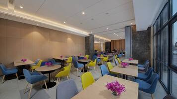 A modern dining area with multiple tables and chairs, a kitchen in the background, and large windows.