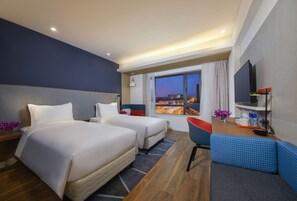 A hotel room with two beds, a desk, a chair, a TV, and a large window offering a city view.