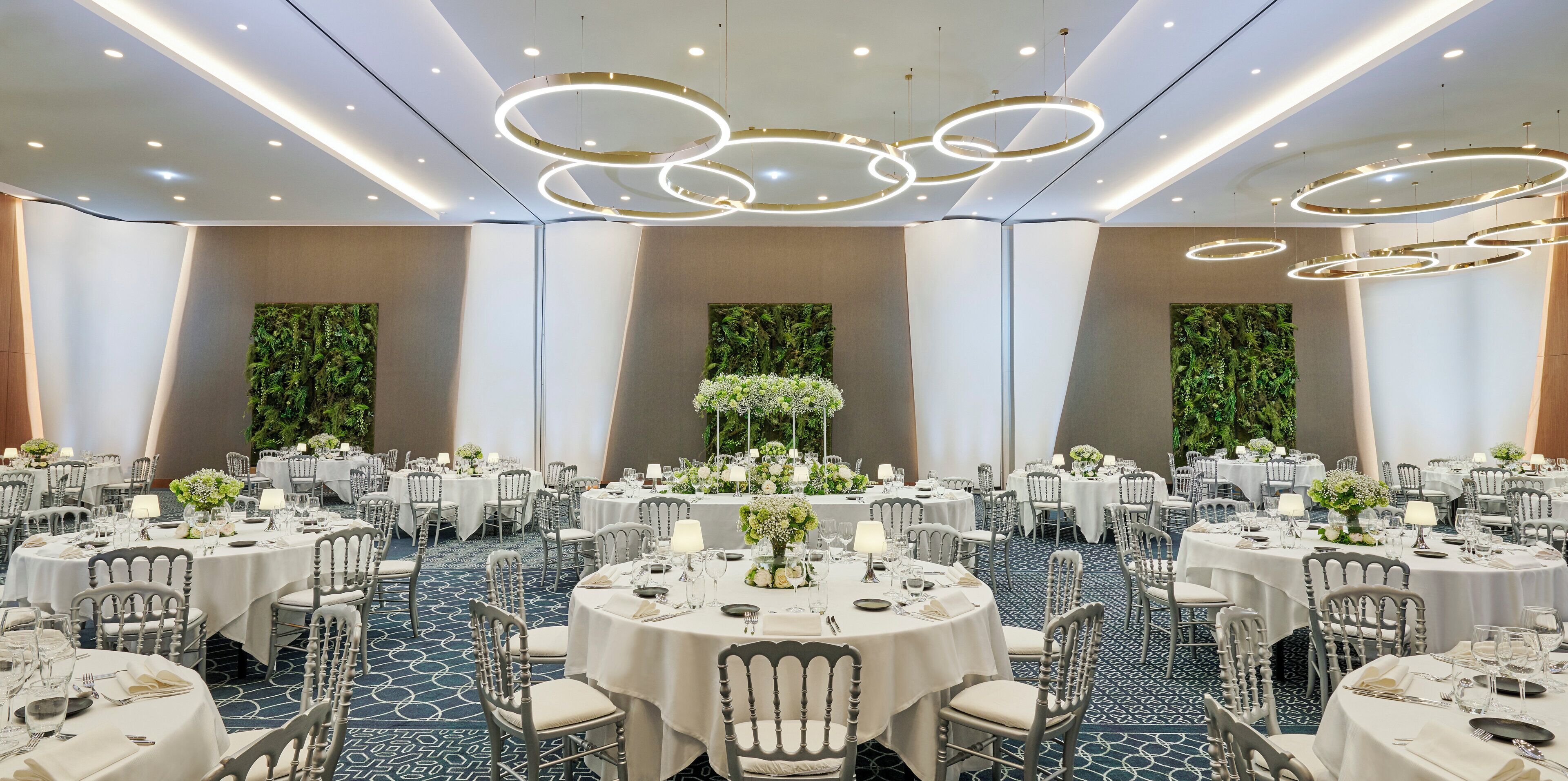 A banquet hall with round tables, white tablecloths, and chairs, featuring a modern ceiling design with circular light fixtures.