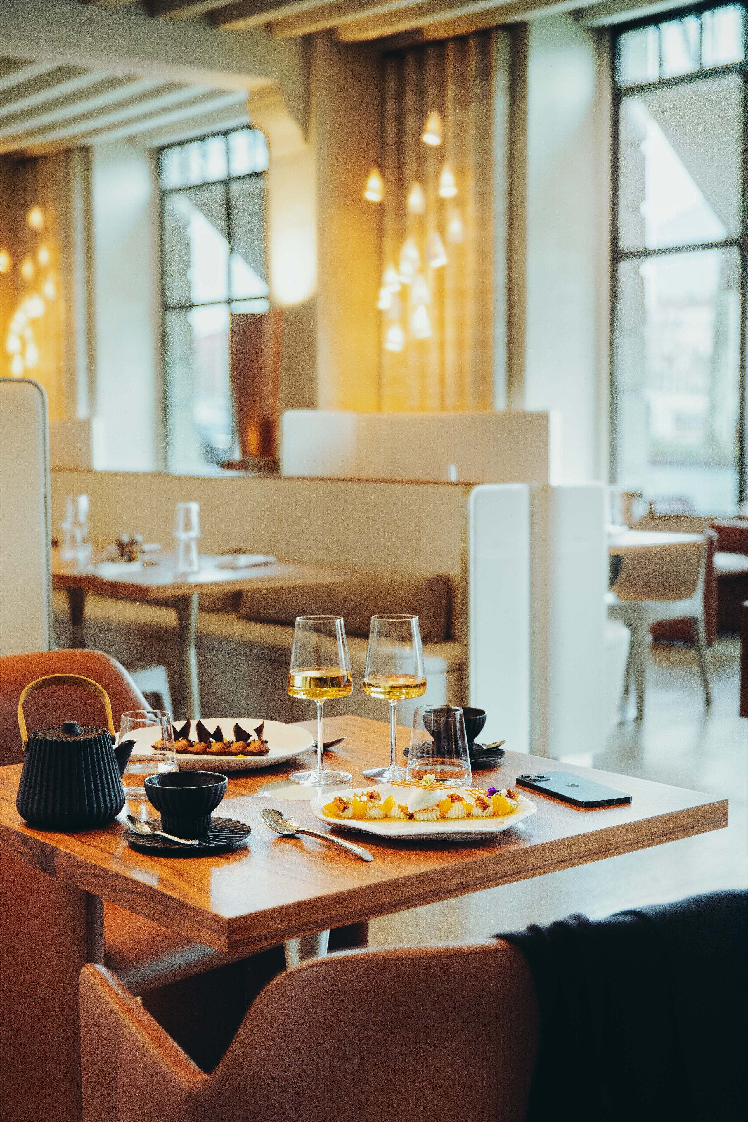 A restaurant setting with a wooden table set for two, featuring a dessert plate, wine glasses, and a smartphone on the table.