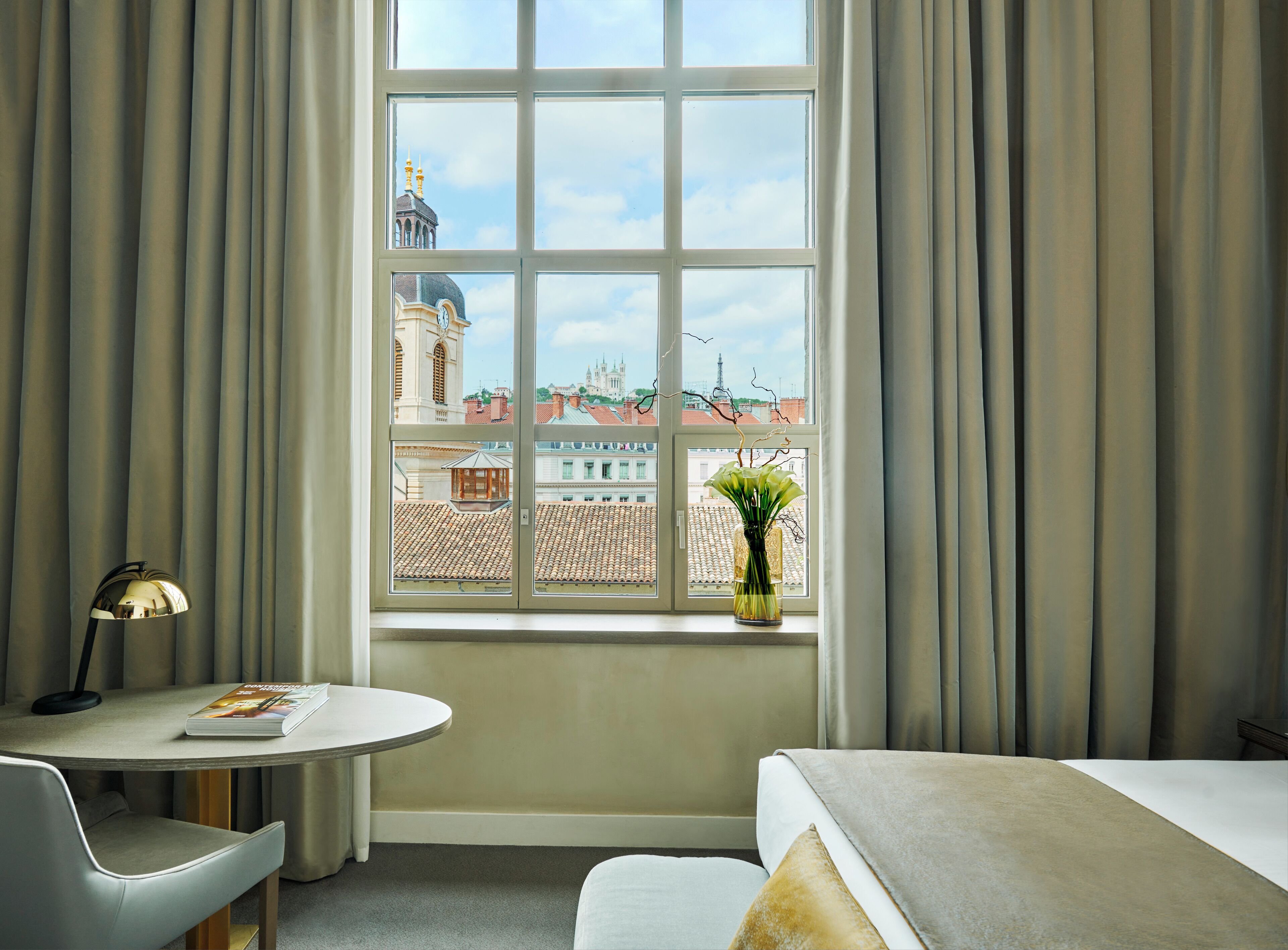 Una habitación de hotel con un ventanal grande, una mesa redonda con una lámpara, una silla, una cama y un jarrón con flores.