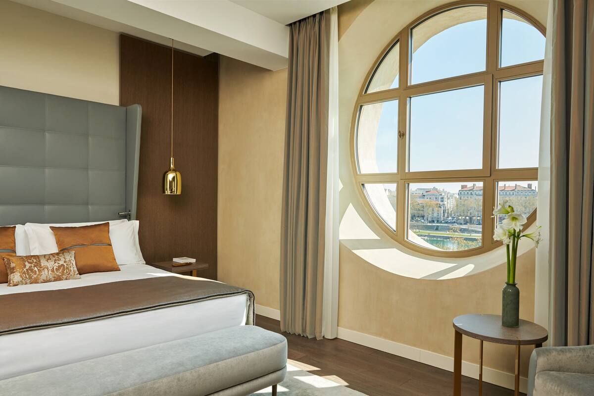 A modern hotel room with a large bed, a round window with a view, a small table with a vase, and a hanging light fixture.