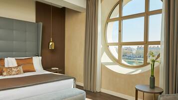 A modern hotel room with a large bed, a round window with a view, a small table with a vase, and a hanging light fixture.