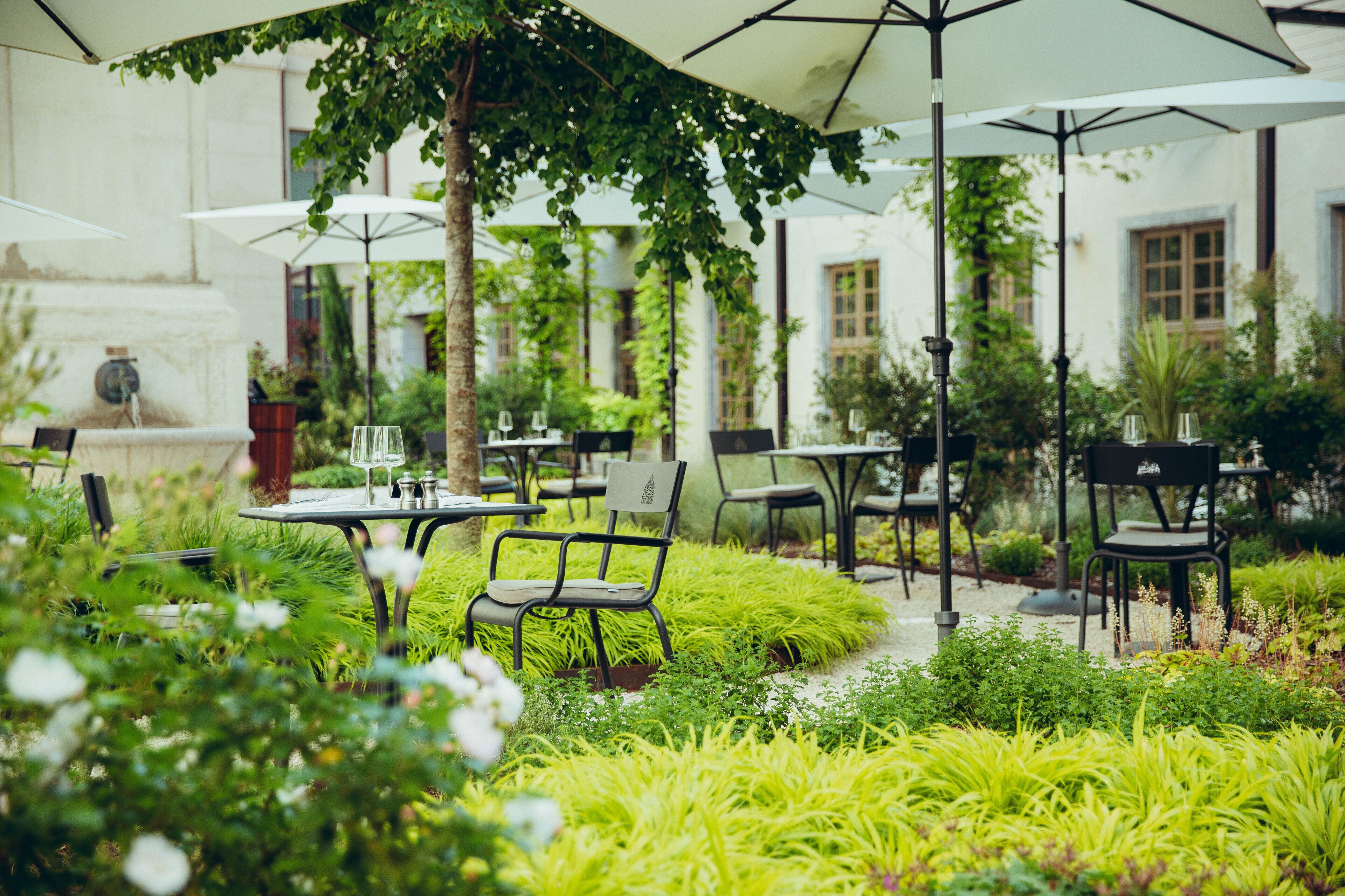 An outdoor cafe with tables and chairs, surrounded by lush greenery and a white building in the background.