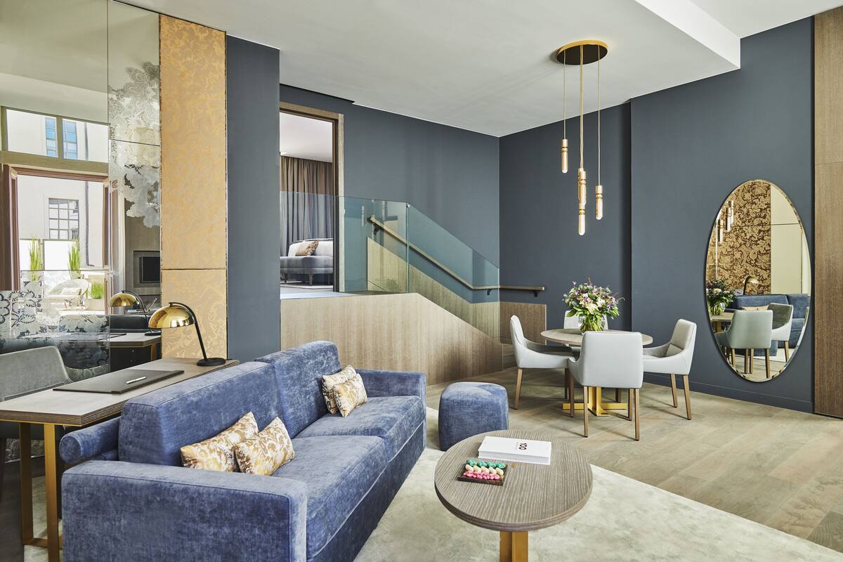 A modern living room with a blue sofa, a dining area with a round table, and a staircase leading to an upper level.