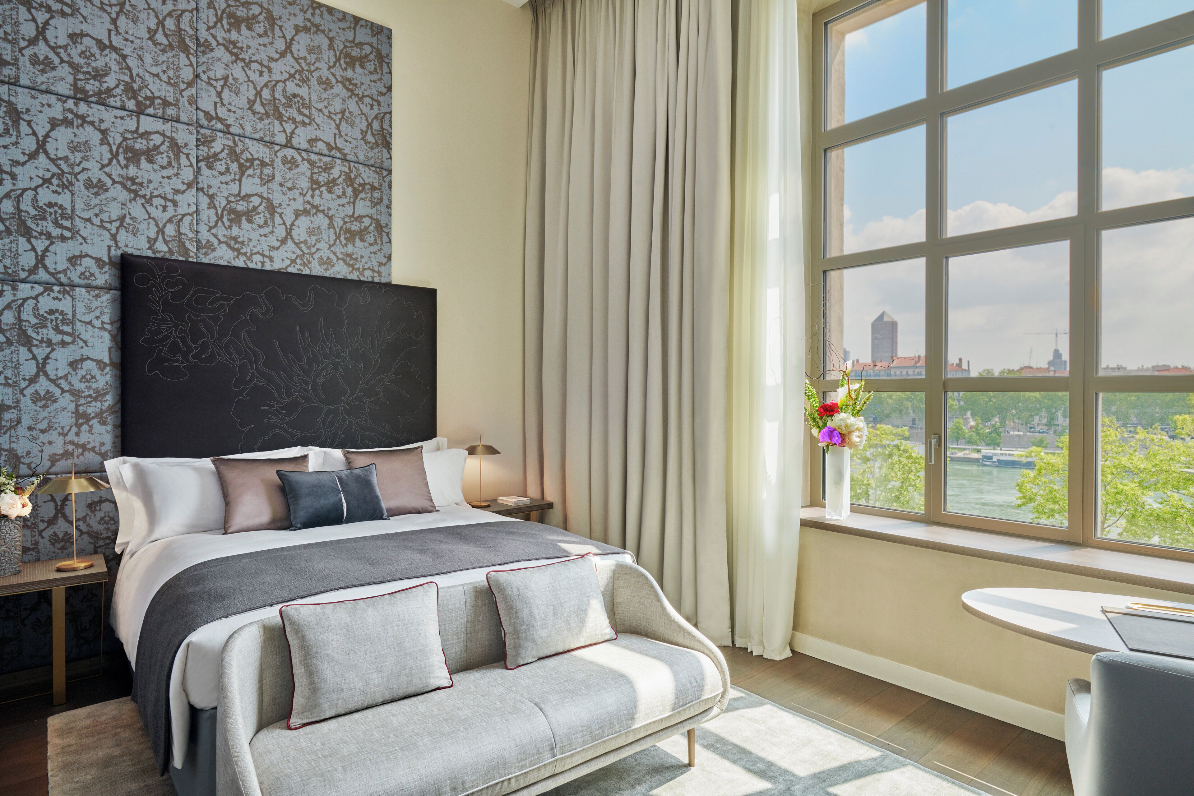Una habitación de hotel moderna con una cama grande, vistas a la ciudad, una ventana con cortinas y una pequeña mesa junto a la ventana.