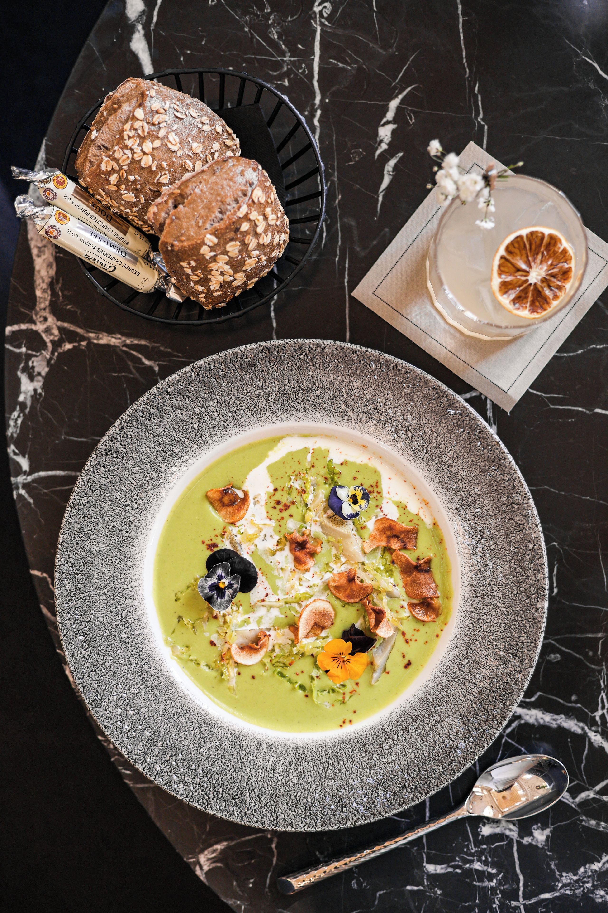 Une assiette de soupe verte, agrémentée de quelques garnitures, une miche de pain et un verre de boisson, le tout posé sur une table en marbre.