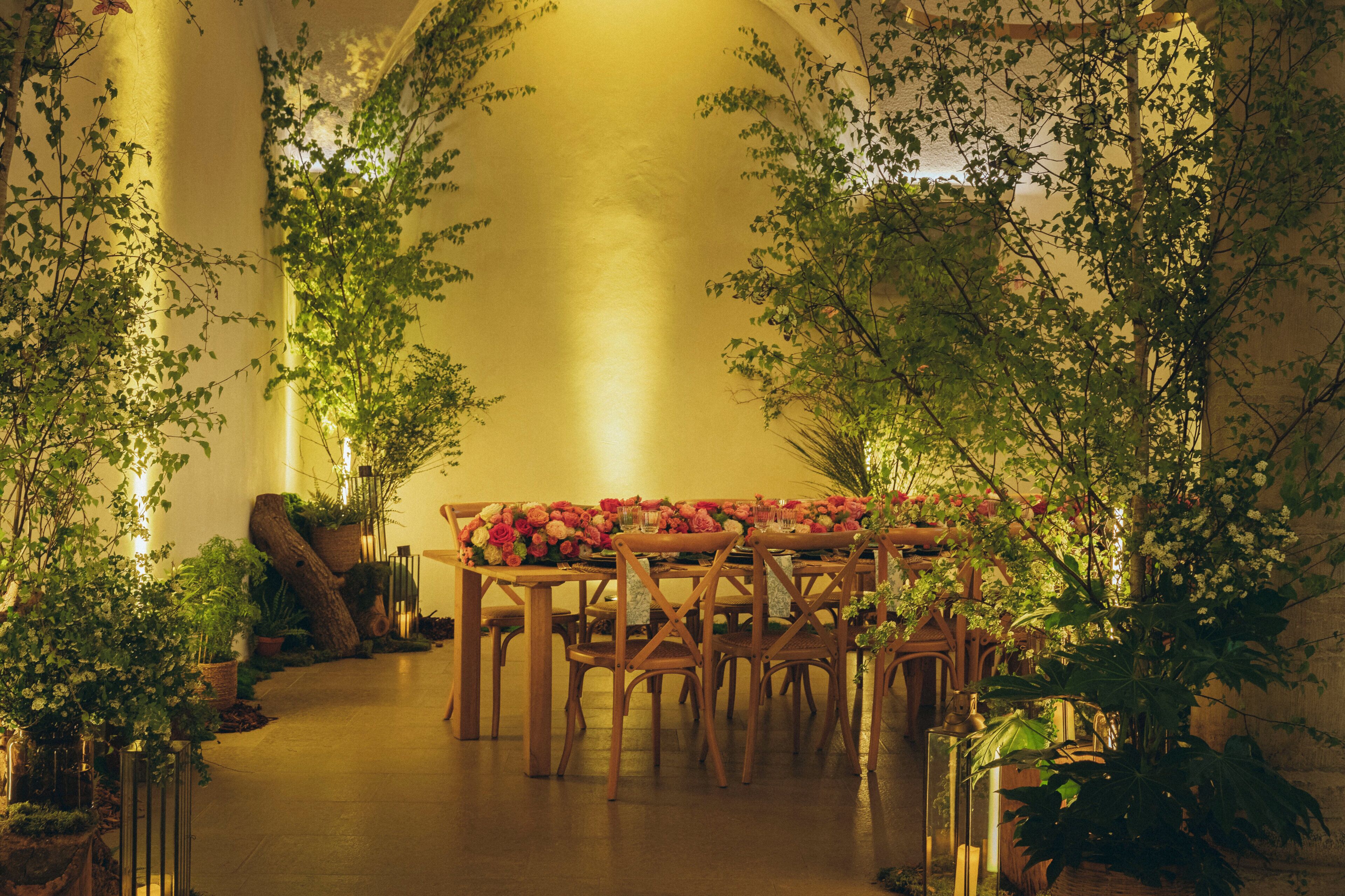 An indoor setting with a long table set for dining, surrounded by potted plants and wooden chairs.