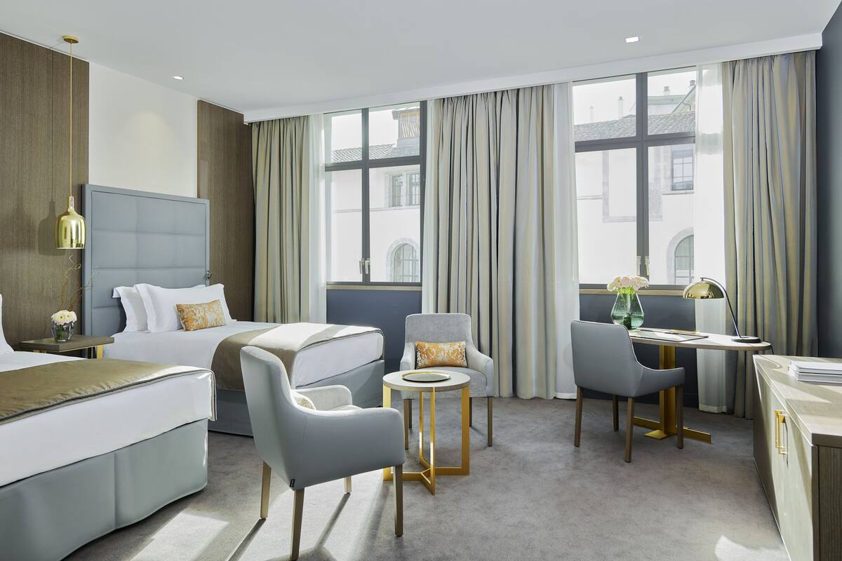 A hotel room with a bed, two armchairs, a small round table, a desk, and a large window with curtains.