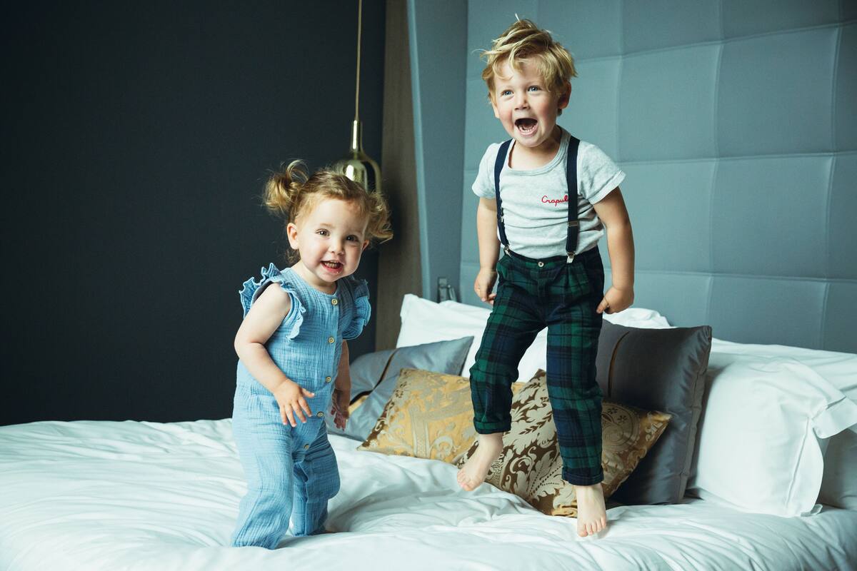 Two children jumping on a bed with a modern headboard and patterned pillows.