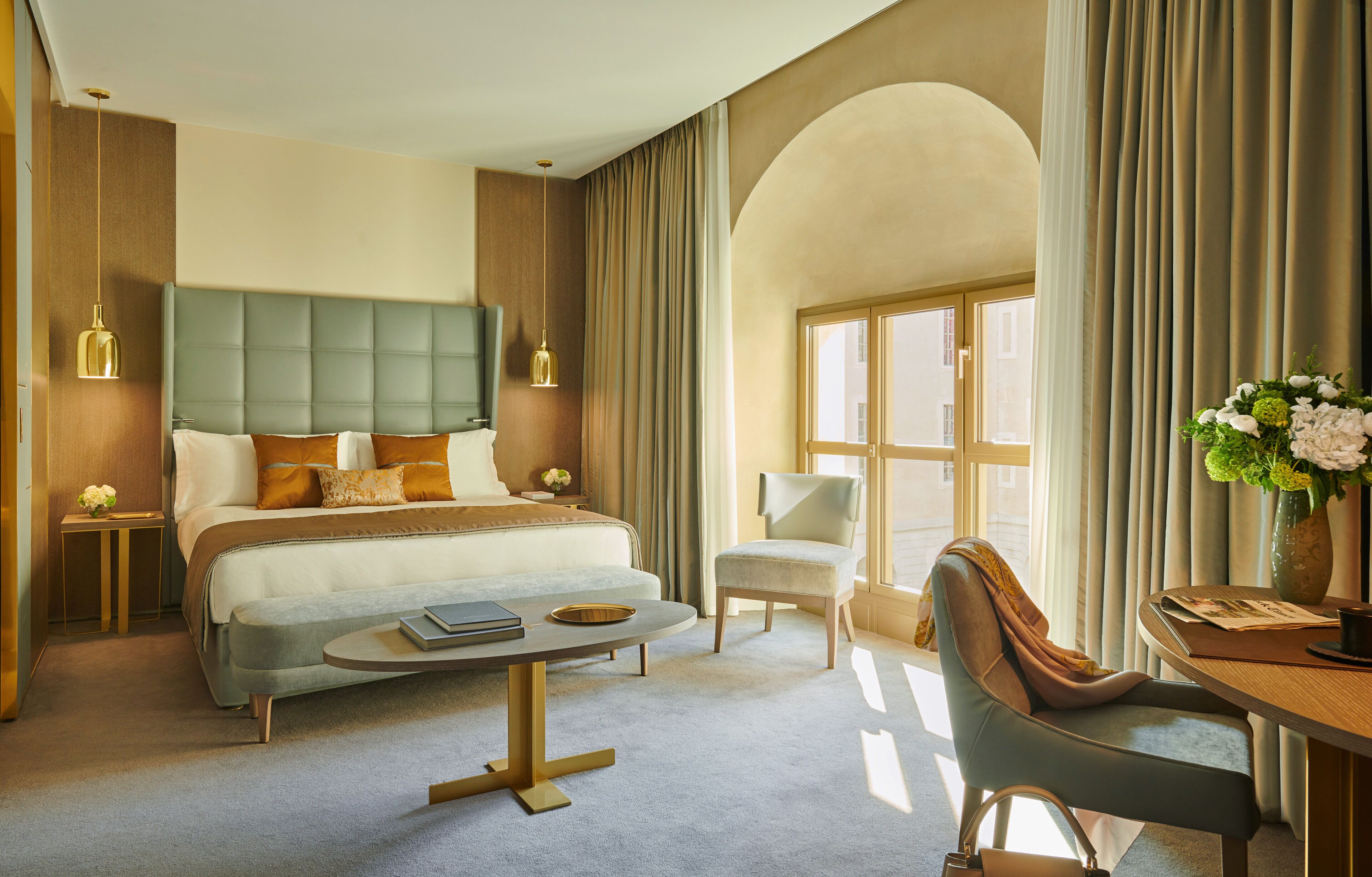 Camera d'albergo con un letto grande, un'area salotto con tavolo e sedie, una scrivania con un vaso di fiori e vista attraverso una finestra con tende.