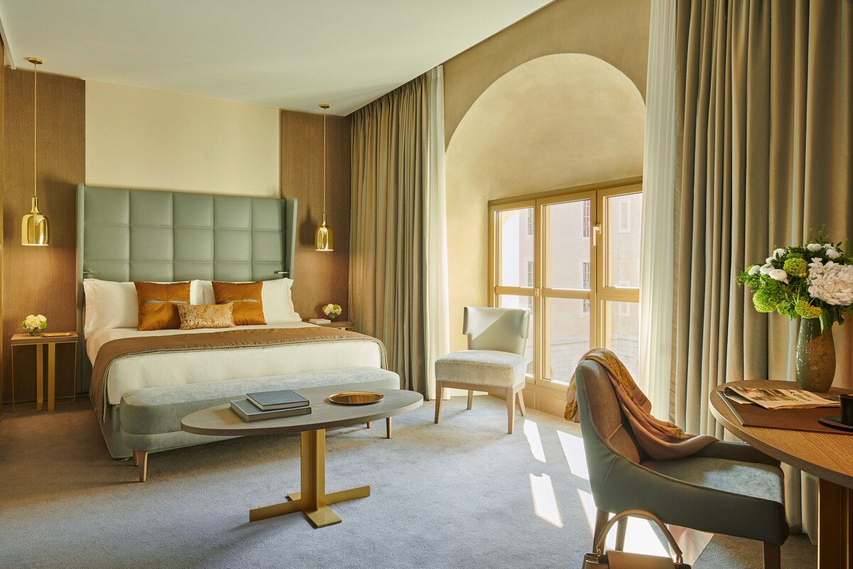 A hotel room with a large bed, a sitting area with a table and chairs, a desk with a vase of flowers, and a view through a window with curtains.
