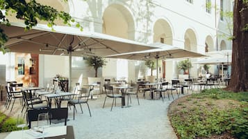 An outdoor café with tables and chairs, surrounded by a garden and a building with arches.
