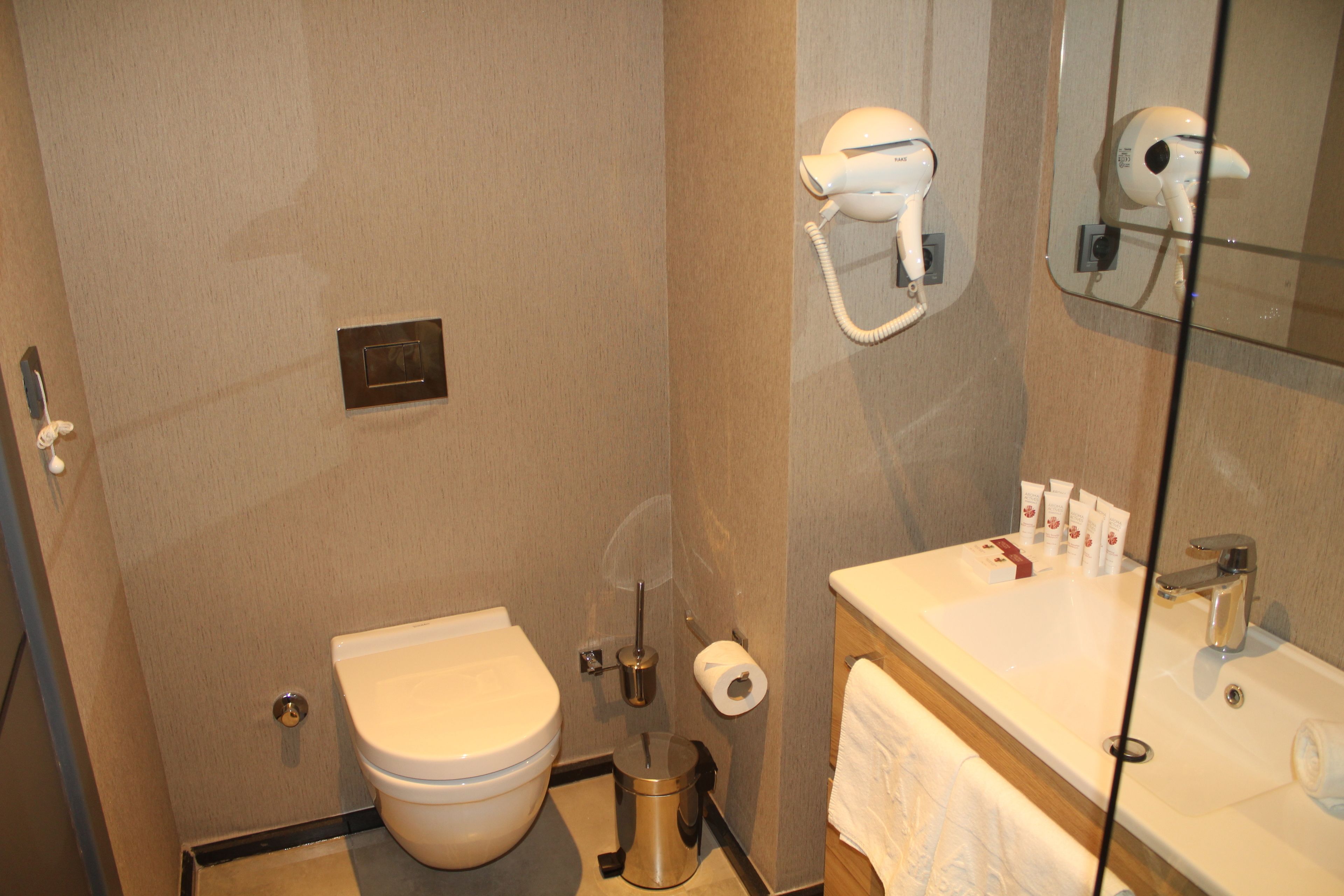 A bathroom with a toilet, bidet, hand dryer, and a sink with toiletries.